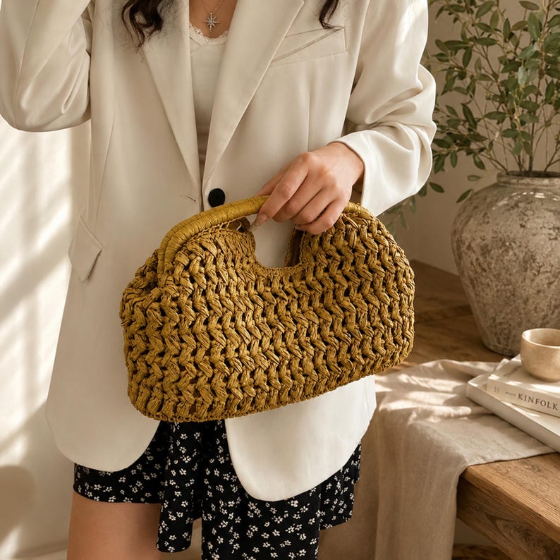 May include: A woven straw handbag with a curved handle. The bag is a golden-yellow color and has a textured, open weave design. The handle is a lighter shade of yellow. The bag is held by a person wearing a white blazer and black and white shorts.