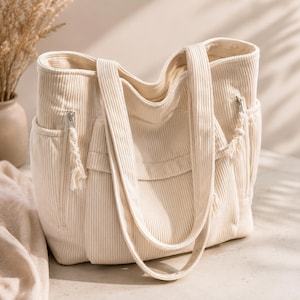 Può includere: Borsa tote in velluto a coste color crema con due tasche esterne, chiusura con cerniera e lunghe tracolle. La borsa ha un aspetto morbido e strutturato e una palette di colori neutri, adatta all'uso quotidiano.