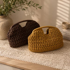 May include: Two handmade crochet handbags. One is dark brown with a matching handle, and the other is mustard yellow with a light brown handle. Both bags have a rounded shape and a woven texture, perfect for carrying essentials.