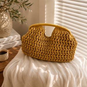 May include: Mustard yellow crochet handbag with a rounded shape and a matching handle. The bag has an open weave design, allowing glimpses of the interior. The handle is a solid, smooth material. The bag is sitting on a white cloth.