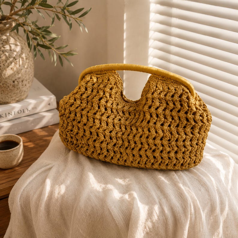 May include: Mustard yellow crochet handbag with a rounded shape and a matching handle. The bag has an open weave design, allowing glimpses of the interior. The handle is a solid, smooth material. The bag is sitting on a white cloth.