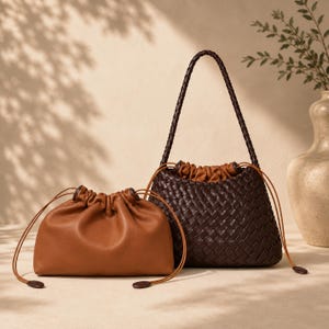 May include: Two leather bags against a neutral backdrop. A small, tan drawstring pouch is in front of a larger, dark brown woven bag with a braided handle. Both bags feature drawstring closures. The bags are in a studio setting.