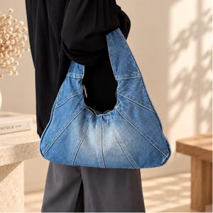 Op de afbeelding: Een blauwe denim hobo-tas in een halvemaanvorm met een patchworkontwerp van gestikte panelen. De tas heeft een brede geïntegreerde schouderband en een zilverkleurige metalen ritssluiting aan de bovenrand, met een vervaagde wassing.