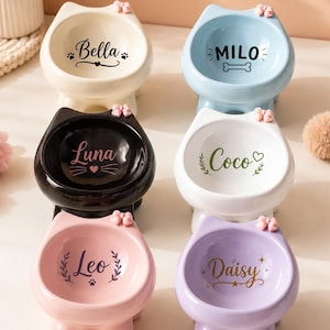 Pode incluir: Seis taças de cerâmica em forma de cabeça de gato com pequenos laços cor-de-rosa nas orelhas. As taças são creme, azul-claro, pretas, brancas, cor-de-rosa e lavanda, cada uma com um nome personalizado: Bella, Milo, Luna, Coco, Leo e Daisy com ícones.
