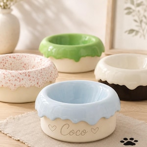 Puede incluir: Cuatro cuencos de cerámica para mascotas con forma de dónut y glaseados en azul claro, verde, rosa moteado y blanco con bases oscuras. El cuenco azul claro tiene el nombre Coco grabado en el lateral entre dos pequeños símbolos de corazón.