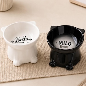 Pode incluir: Dois recipientes de cerâmica em forma de cabeça de gato, um branco e um preto. O recipiente branco apresenta o nome Bella com pegadas, e o preto apresenta o nome Milo com um gráfico de osso. Ambos têm pequenas orelhas e pés.