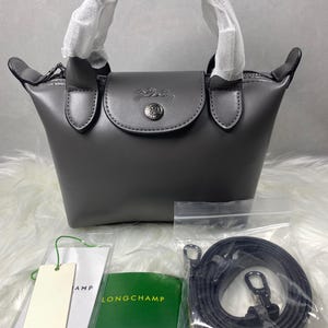 Pode incluir: Uma bolsa cinza com uma alça superior e fecho de aba. A bolsa tem um emblema prateado na frente e vem com uma alça de ombro removível. Inclui etiquetas e um cartão verde com o nome da marca.