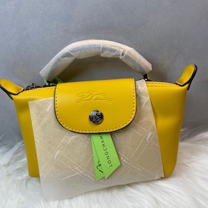 May include: A bright yellow handbag with a flap closure and a top handle. The bag features a logo of a running horse on the flap. A green tag with the brand name is attached. The bag is sitting on a white, fluffy surface.