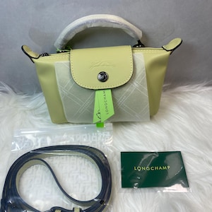 May include: A pale yellow handbag with a top handle and a flap closure. The bag features a silver-tone button and a small embossed logo. A detachable navy blue strap and a green tag are included. The brand name "LONGCHAMP" is printed on a green card.