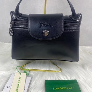 May include: Black leather handbag with a flap closure and a zipper pocket. The bag features a silver-tone logo emblem and a small handle. The brand name "LONGCHAMP" is visible on the tag and card.