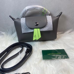 May include: A gray handbag with a top handle and a detachable shoulder strap. The bag has a flap closure and a green tag that says "LONGCHAMP". The bag is sitting on a white surface.