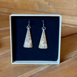 Op de afbeelding: Een paar koperkleurige, driehoekige oorbellen met een wit rendierontwerp, gepresenteerd in een witte doos met een zwart interieur. De oorbellen hangen aan zilveren haken. De doos staat op een houten oppervlak.