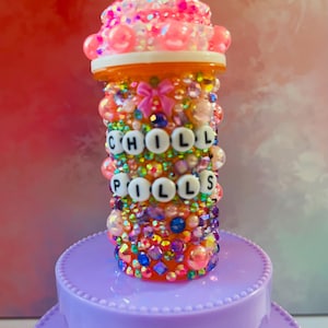 May include: A decorative pill bottle covered in colorful beads, gems, and pearls. The bottle has the words "CHILL PILLS" spelled out in white letter beads. The top is adorned with pink and clear beads and a small pink bow.