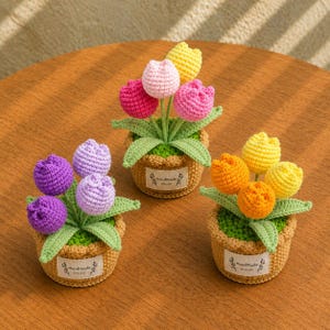Peut inclure: Trois plantes en crochet en pot avec des fleurs en forme de tulipes aux couleurs violet, rose, jaune et orange. Chaque plante est dans un pot en crochet marron avec une étiquette en tissu blanc portant l'inscription Handmade with love.