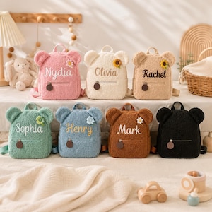 Peut inclure: Collection de huit sacs à dos en peluche en forme d'ours, de différentes couleurs : rose, crème, beige, vert, bleu, marron et noir. Chaque sac à dos porte un nom brodé sur le devant et une fleur décorative. Ils ont une petite poche avant et des bretelles réglables.