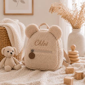 Peut inclure: Un sac à dos beige en forme d'ours en peluche avec le nom "Chloe" brodé sur le devant. Le sac à dos a une fermeture éclair et des bretelles réglables. Un petit ours en peluche crocheté et des blocs de bois sont à proximité.