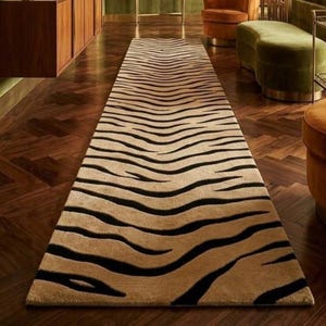 May include: A long, rectangular runner rug featuring a repeating black tiger stripe pattern set against a tan, textured background. The rug is positioned centrally on a dark brown wooden floor with a herringbone pattern, extending into the distance.
