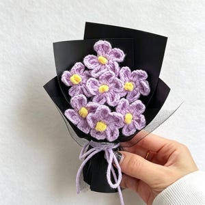 Könnte beinhalten: Ein kleiner Blumenstrauß aus sechs gehäkelten Blüten mit hellvioletten Blättern und leuchtend gelben Mitten. Die Blumen sind vor einem schwarzen Papierhintergrund angeordnet, in schwarzes Netzgewebe eingewickelt und mit einer hellvioletten Garnschleife gebunden.