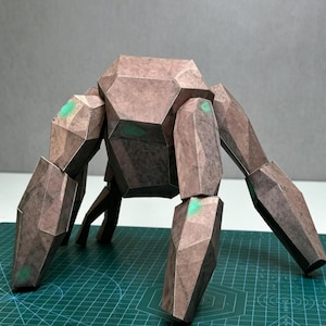 Könnte beinhalten: Eine geometrische Papierbastelfigur mit einer braunen, texturierten, steinartigen Oberfläche. Die Kreatur hat einen massigen Rumpf und vier lange, kantige Gliedmaßen. Kleine, leuchtend grüne kreisförmige Akzente sind an den Gelenken sichtbar.