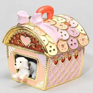 Könnte beinhalten: Eine dekorative Schmuckdose aus Metall in Form einer Hundehütte mit aufklappbarem Dach. Die Hütte ist in Pastellrosa und Creme gehalten, mit Goldverzierungen, einer hellrosa Schleife oben und einer kleinen weißen Hundefigur im Eingang.