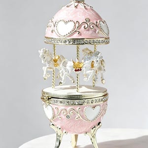 Könnte beinhalten: Eine rosa-goldene Schmuckdose im Karussell-Design mit weißen Pferden zwischen einer Kuppel und einem runden Sockel. Das Äußere ist mit weißen herzförmigen Akzenten, klaren Kristallen und goldenen Verzierungen versehen und steht auf drei geschwungenen Beinen.