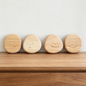 Könnte beinhalten: Vier helle Holzobjekte mit glatten, abgerundeten, kieselsteinartigen Formen. Jedes Stück zeigt ein einfaches, eingraviertes Gesicht mit zwei kleinen Punkten als Augen und einer geschwungenen Linie als Mund, mit sichtbarer Holzmaserung.