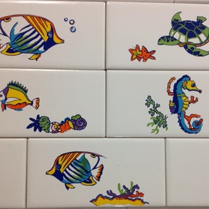 May include: White ceramic tiles with colorful sea life illustrations. The designs include fish, a sea turtle, a starfish, a seahorse, and coral. The artwork features bright colors and playful designs, suitable for a bathroom or kitchen.