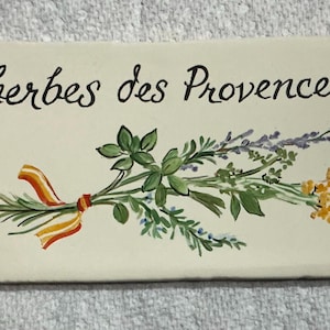 May include: A rectangular off-white ceramic tile featuring the black cursive text herbes des Provence. The center displays a hand-painted illustration of a bundle of green herbs tied with a yellow and orange ribbon, with small blue corner accents.