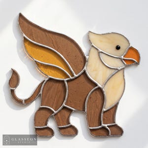 May include: A stained glass griffin suncatcher featuring brown, cream, and amber glass segments joined by silver solder lines. The mythical creature has a lion-like body, eagle wings, and a beak. The text GLASSEON STUDIO is in the corner.