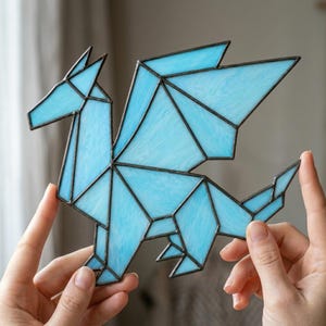 Könnte beinhalten: Ein Drachen-Sonnenfänger aus Buntglas mit hellblauen, halbtransparenten Glasscheiben, die durch dunkle Metalllötlinien verbunden sind. Der Drache ist in einer geometrischen, stilisierten Form mit spitzem Kopf, großen Flügeln und einem Schwanz dargestellt.