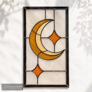 May include: A rectangular stained glass panel featuring a crescent moon and two four-pointed stars in shades of amber and gold. The design is set against a textured, translucent white glass background, all encased within a dark metal frame.