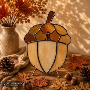 May include: Stained glass acorn with a warm color palette of amber, brown, and cream. The decorative piece is displayed on a small gold stand, surrounded by fall leaves, acorns, and a vase of autumn foliage. The image includes the "GLASSEON STUDIO" logo.