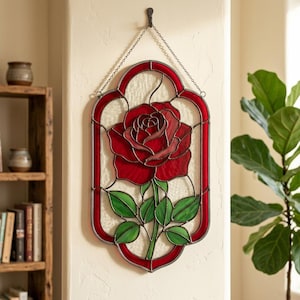 Puede incluir: Un colgante de pared de vitral que presenta una rosa roja central con hojas verdes, encerrada en un borde rojo con un fondo blanco texturizado. La pieza está suspendida por una cadena de metal de un gancho, mostrando una intrincada artesanía.