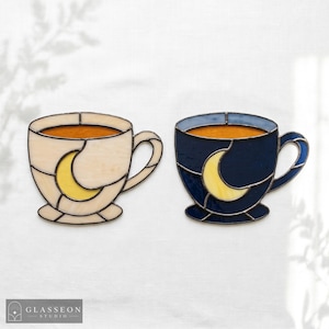 Op de afbeelding: Twee glas-in-lood koffiekopjes. Eén is crèmekleurig met een gele halve maan, de andere is marineblauw met een gele halve maan. Beide kopjes hebben een bruine vloeistof.