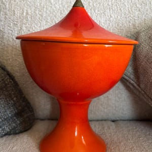 May include: A vibrant orange ceramic lidded jar with a pedestal base. The jar has a rounded body and a conical lid, topped with a bronze-colored finial. The glossy finish reflects light, highlighting the bright color.