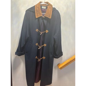 May include: A long, dark navy blue coat featuring a tan suede collar with decorative stitching. The coat includes three toggle closures with leather loops and wooden toggles down the front, long sleeves with folded cuffs, and a dark purple inner lining.