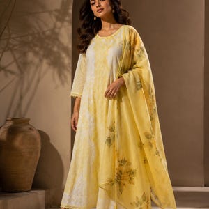 May include: A long-sleeved, floor-length dress in white and yellow with a subtle tie-dye pattern and scalloped hem details. The outfit includes a matching sheer yellow dupatta featuring a delicate floral print in muted green and yellow tones.