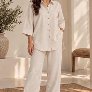 Può includere: Un completo beige a due pezzi composto da una camicia abbottonata con maniche a tre quarti e orlo arrotondato, abbinata a pantaloni a gamba larga. L'outfit presenta un tessuto testurizzato ed è indossato con sandali chiari aperti.