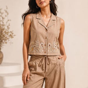 May include: A sleeveless tan collared vest with a button-down front and white floral embroidery along the hem, paired with matching tan wide-leg trousers featuring a drawstring waist and coordinating floral embroidery near the bottom hem.