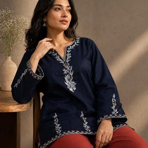 Könnte beinhalten: Eine dunkelblaue langärmelige Tunika mit geschlitztem Ausschnitt und aufwendiger Blumenstickerei in Weiß-, Hellblau- und Brauntönen entlang des Kragens, der Ärmelbündchen und des Saums, kombiniert mit einer rostfarbenen Hose.