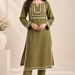 May include: An olive green long-sleeved tunic and matching trousers set. The tunic features a notched neckline and white decorative trim with colorful zigzag stitching along the chest, cuffs, and hem. The trousers have matching trim at the ankles.