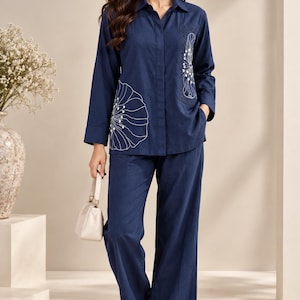 Peut inclure: Un ensemble bleu marine composé d'une chemise à manches longues boutonnée avec des broderies florales blanches et d'un pantalon large assorti. La chemise présente un col et une poche poitrine. Un petit sac à main crème et des sandales crème.