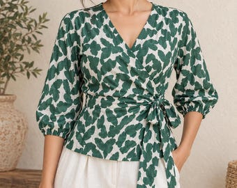 May include: A wrap-style blouse featuring a V-neckline, three-quarter length puff sleeves with elastic cuffs, and a side-tie waist closure. The garment is made of a white fabric with a repeating dark green abstract leaf or petal pattern.