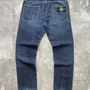Op de afbeelding: Donkerblauwe spijkerbroek van denim met rechte pijpen, voorzien van een wit merketiket op de achterband en een rechthoekige zwarte patch met een geel en groen kompaslogo op de rechter achterzak, met subtiele vervaagde wassingdetails.