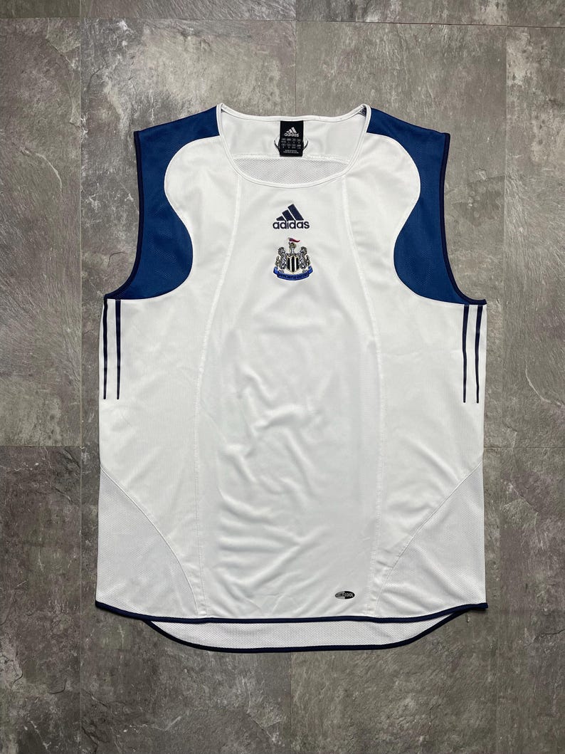 Puede incluir: Camiseta de f&uacute;tbol blanca sin mangas con detalles azul marino en hombros y laterales. Presenta el logo de Adidas y el escudo del equipo en el pecho. Tres rayas azul marino a cada lado.