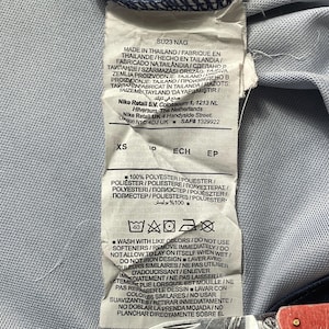 May include: A white fabric garment care label with black text, indicating the item is made in Thailand, size XS, and composed of 100% polyester. The label includes washing instructions, symbols, and the text SU23 NAG, Nike Retail B.V., and Nike Retail UK.