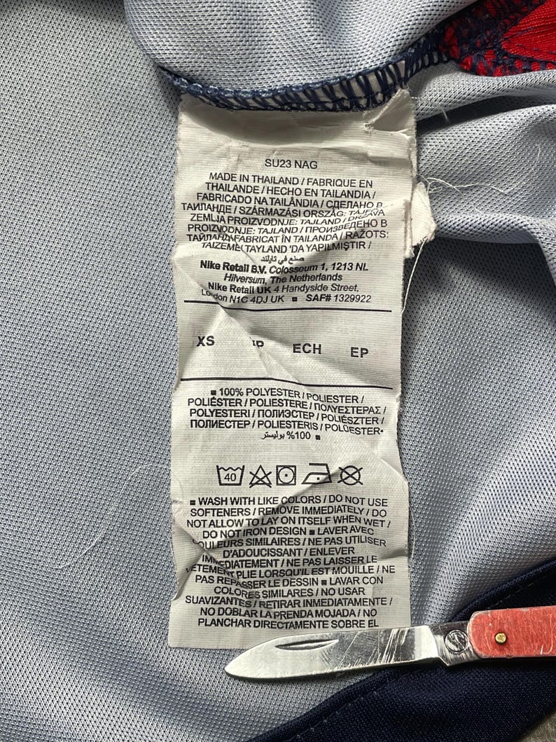 May include: A white fabric garment care label with black text, indicating the item is made in Thailand, size XS, and composed of 100% polyester. The label includes washing instructions, symbols, and the text SU23 NAG, Nike Retail B.V., and Nike Retail UK.