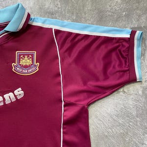 May include: Maroon and light blue West Ham United football shirt. The shirt displays the club crest with a shield and the words "West Ham United". The collar and sleeve cuffs are light blue with white stripes. White piping runs down the sides.