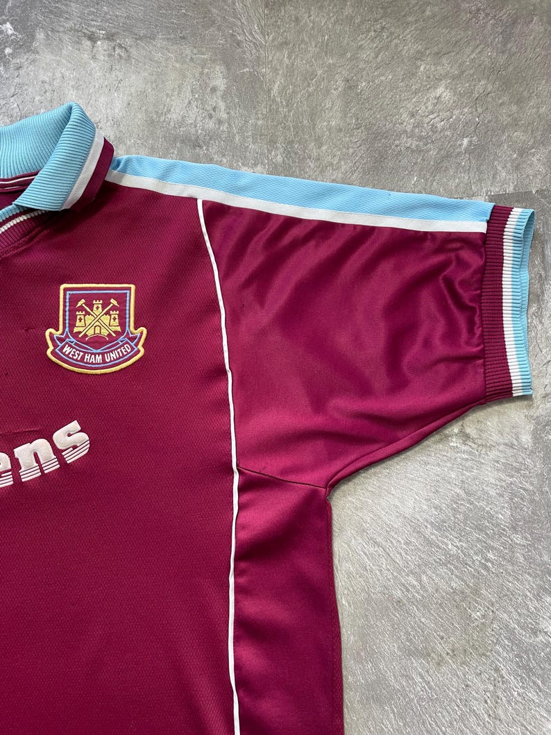 May include: Maroon and light blue West Ham United football shirt. The shirt displays the club crest with a shield and the words "West Ham United". The collar and sleeve cuffs are light blue with white stripes. White piping runs down the sides.
