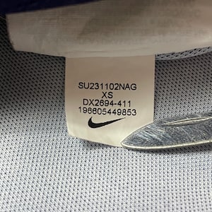 May include: A white clothing label sewn onto a light blue textured fabric. The label displays the text SU231102NAG, XS, DX2694-411, 196605449853, and a black Nike swoosh logo. A metallic object is partially visible at the bottom right of the label.